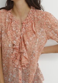 Peach floral blouse with ruffled neckline and button closure. Sheer fabric with a delicate pattern of small white flowers. Short sleeves.
