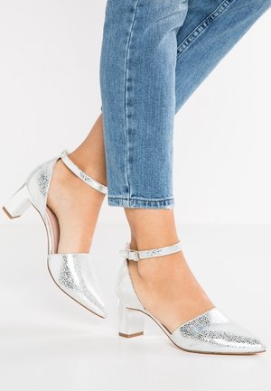 Personne portant des chaussures argentées à talon bas épais, avec des brides à la cheville et un jean bleu retroussé sur fond blanc.