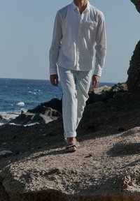 Homme portant une chemise blanche, un jean clair et des sandales marchant sur un terrain rocheux près de l'océan sous un ciel clair.