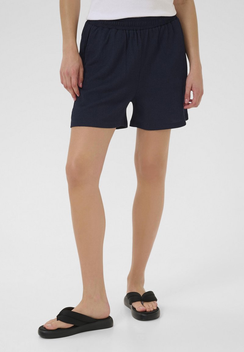 Shorts bleu marine avec une ceinture élastique, texture douce et finition lisse, associés à des sandales flip-flop noires avec un large design de lanière.