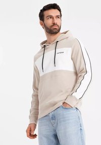 Beige a bílá mikina s černými akcenty a stahovacími šňůrkami, se znakem na hrudi. Měkká tkanina, volný střih a klokánková kapsa.
