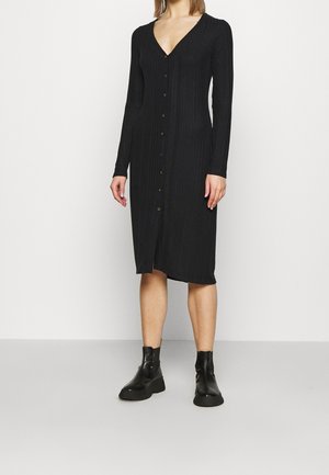 Femme portant une robe midi noire côtelée avec boutons, manches longues et bottines noires, debout devant un fond blanc uni.