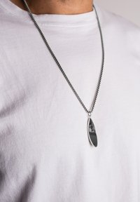 Colier din lanț argintiu cu un pandantiv alungit, oval, având o suprafață neagră lustruită, purtat peste un tricou alb simplu.