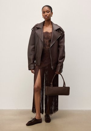 Femeie purtând o jachetă maro închis din piele supradimensionată, rochie transparentă din dantelă cu franjuri deasupra coapselor, purtând o geantă maro asortată.