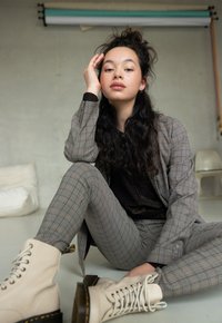 Beige gevechtslaarzen met veters, grijs geruiten pak, slim-fit broek, gestructureerd zwart topje, zittende pose, binnenomgeving met minimale achtergronddetails.