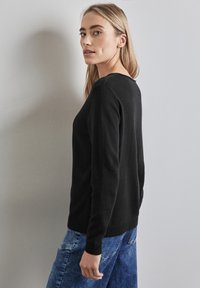 Schwarzer Pullover mit langen Ärmeln, Rundhalsausschnitt und gerippten Bündchen. Der Stoff wirkt glatt und eng anliegend, ergänzt durch blaue Denim-Hosen.
