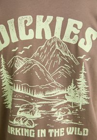 Barna pamut póló, amelyen egy világoszöld grafikán hegyek, fák és sziklák láthatók a "DICKIES WORKING IN THE WILD" felirattal.