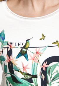 Kortærmet hvid t-shirt med et farverigt print af kolibrier og blomster, med grønne blade og et sort geometrisk mønster.
