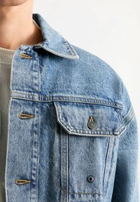 Hellblaues Jeansjacke mit klassischem Kragen, Knopfakzenten und einer Brusttasche mit markierter Hardware und kontrastierenden Nähten.
