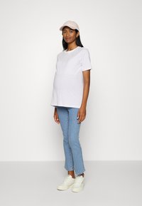 Top de maternidade em branco com riscas lilás claras, gola redonda, mangas curtas. Combinado com jeans azul claro e ténis brancos. Boné rosa usado.