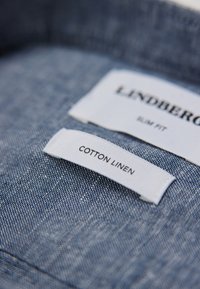 Smal passform skjorta i blått bomull-lin-blandning. Taggar visar varumärket "Lindberg" och information om tyget. Synlig texturerad tygmönster.