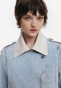 Chaqueta de mezclilla con un lavado azul claro, gran cuello beige, detalles de botones y costuras en contraste. Presenta bolsillos laterales y un cierre de cremallera frontal.