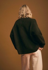 Übergroßer grüner Pullover aus weichem Material mit weiten Ärmeln, kombiniert mit lässigen, hellrosa Jeans im Relaxed-Fit, vor einem braunen Hintergrund.