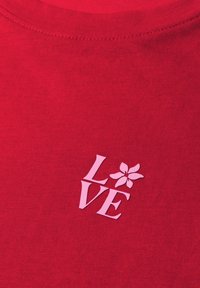 Czerwona tkanina z różowym napisem "LOVE", gdzie kwiat zastępuje literę "O" na wysokości klatki piersiowej ubrania.