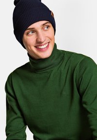 Zielony sweter z golfem w prążki z małym logo na piersi, zestawiony z granatową wełnianą czapką z tanowym skórzanym akcentem.