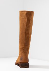 Stivale in suede marrone alto con tacco in legno basso e impilato, cuciture visibili e un tenue logo a forma di farfalla vicino al tacco, mostrato da dietro.