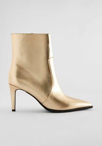 Kultainen metallinen nilkkuri, jossa on terävä kärki ja ohut stiletto-korko. Tasainen pinta, minimalismin design ja sivuommelu-yksityiskohta.