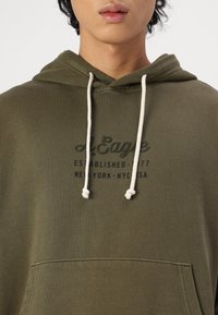 Oliven grønn hettegenser laget av bomull, med en frontlomme, snøring og trykt tekst i svart og mørkegrønn.