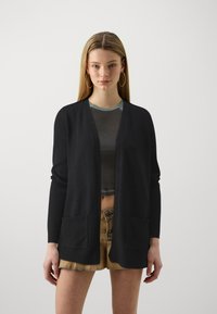Svart cardigan med V-hals-design, som har to frontlommer og ribbet kant. Matchet med en cropped topp og denimshorts.