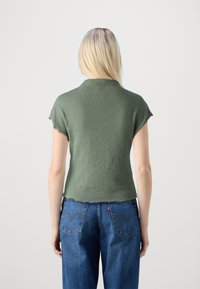 Blondýna stojí zády, má na sobě krátký rukáv zelený pletený top a modré džíny s viditelnými zadními kapsami.