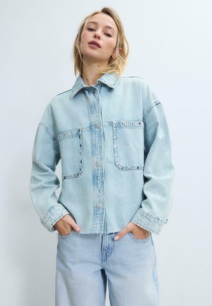 Mlada ženska z rokami v žepih, oblečena v svetlo modro jeans jakno z metalnimi zakovami in ujemajoče se svetlo modre jeans hlače, na enobarvni podlagi.