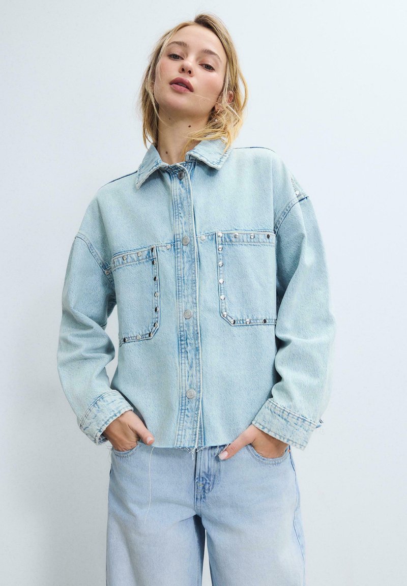Jeune femme les mains dans les poches, portant une veste en denim bleu clair ornée de clous métalliques et un jean assorti bleu clair, sur un fond uni.