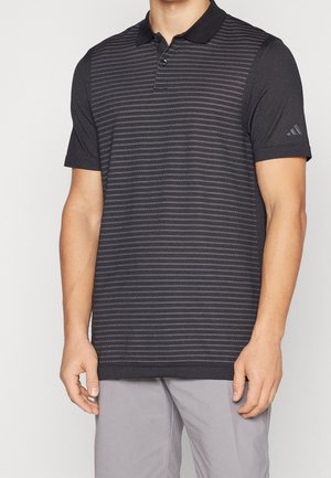 Homme portant un polo à manches courtes noir avec de fines rayures horizontales et un petit logo sur la manche gauche.