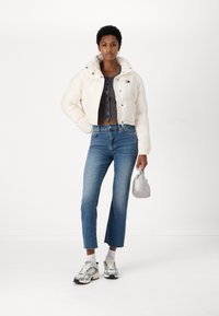 Giacca a vento bianca cropped, camicia grigia con bottoni, jeans blu a gamba dritta e sneakers argento. Piccola borsa argento, in piedi contro uno sfondo semplice.