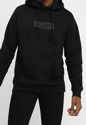 Persoon draagt een zwarte Tommy Jeans hoodie met voorzak en trekkoorden, gecombineerd met een zwarte broek, tegen een effen achtergrond.
