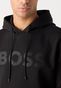 Čierna mikina s kapucňou vyrobená z mäkkej tkaniny, s veľkým tónovým logom "BOSS" na prednej strane a so šnúrkami na zapínanie okolo krku.