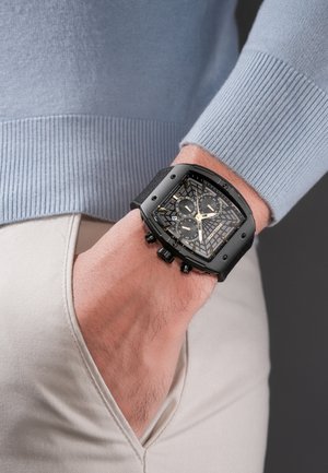 Persoon draagt een zwart vierkant chronograafhorloge met een patroon op de wijzerplaat, gouden wijzers en een zwarte stoffen band over een lichtbeige broekzak.