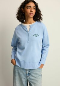 Lyseblå sweatshirt lavet af bomuld, med en henley-udskæring med knapper. Teksten "NEW YORK CITY NY" broderet i grøn. Løst fit.