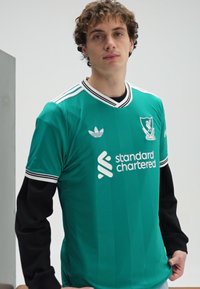 Grønn fotballtrøye laget av lett materiale. Har kontrasterende svarte ermer, hvite striper på kragen og ermene, og en logo.