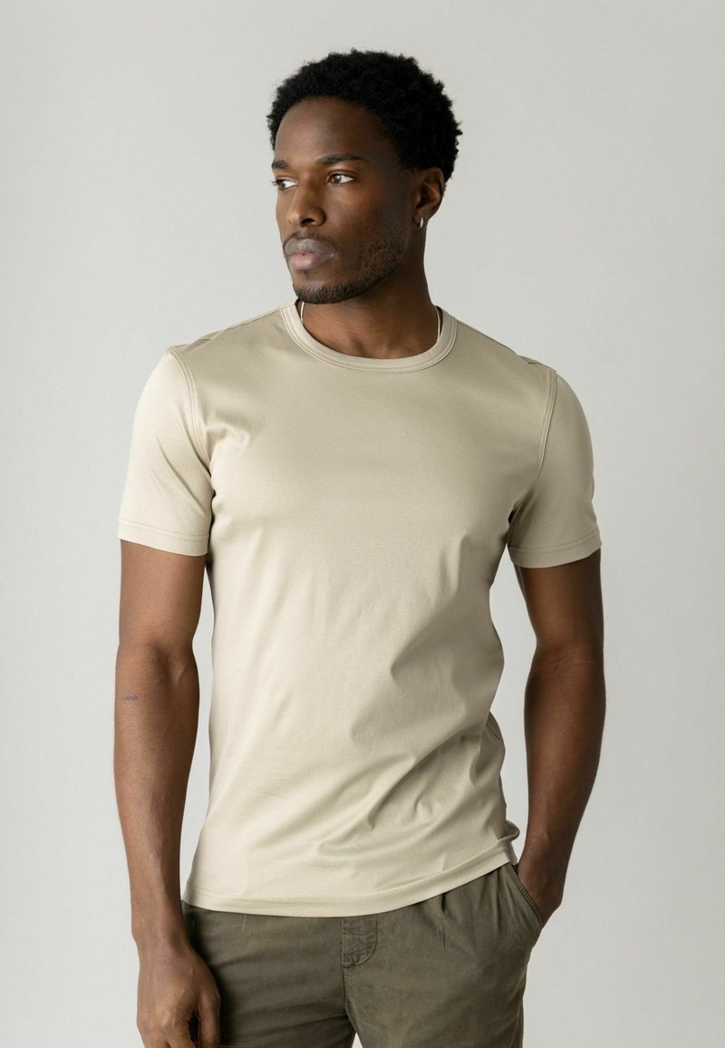 Moški s kratkimi kodrastimi lasmi, oblečen v bež kratko rokavno majico in olivne hlače, gleda v stran z eno roko v žepu.