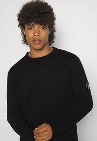 Felpa nera con scollo rotondo, polsini a costine e dettagli di cucitura. Presenta una piccola toppa con logo sul maniche sinistro.