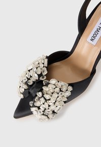 Scarpa con tacco alto in satin nero, caratterizzata da una punta affusolata e un grande fiocco tempestato di cristalli nella parte anteriore, con un interno beige.