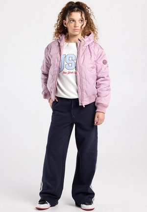 Tienermeisje met krullend haar, een roze doorgestikte jas, een witte sweater met de tekst "USA", marineblauwe wijde broek en sneakers, staand met handen in de zakken.