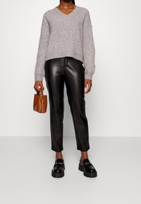 Osoba v šedém pleteném svetru, černých kožených kalhotách, černých mokasínech a s malou hnědou kabelkou, stojící na jednobarevném pozadí.