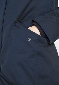 Main dans la poche avant d’une veste bleu marine avec une fermeture à pression sur le rabat de la poche.