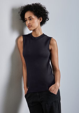 Top nero senza maniche con colletto alto, realizzato in un tessuto liscio ed elastico, dalla forma aderente, abbinato a pantaloni scuri, appoggiato a un muro.