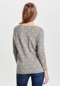 Pull gris avec des pois jaunes. Doté d'un col large et de manches longues. Tissu en maille douce ; coupe décontractée, porté avec un jean bleu.