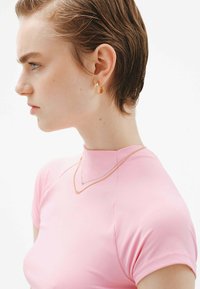 Top corto de manga corta ajustado en color rosa con un escote alto, combinado con pendientes de aro dorados y cadenas doradas superpuestas, sobre un fondo neutro.