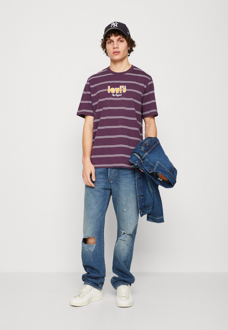 T-shirt violet à rayures avec le logo Levi's, jeans bleus déchirés avec des trous, et une veste en denim à la main. Des baskets blanches et une casquette noire sont portées.