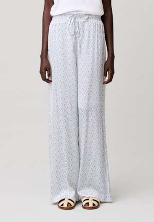 Donna che indossa pantaloni bianchi a gamba larga con piccolo motivo floreale blu e coulisse in vita, abbinati a sandali bianchi con punta aperta.