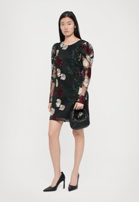 Vrouw in een zwart floral mesh jurk met een zwarte handtas, gecombineerd met zwarte hoge hakken, staande op een witte achtergrond.