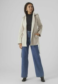 Trench beige clair avec une ceinture à la taille, porté sur un col roulé noir. Assorti avec un jean large taille haute bleu et des bottines noires.