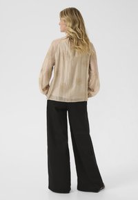 Chemisier en beige transparent avec broderies florales, manches longues et coupe décontractée, associé à un pantalon noir ample. Vue de dos présentée.