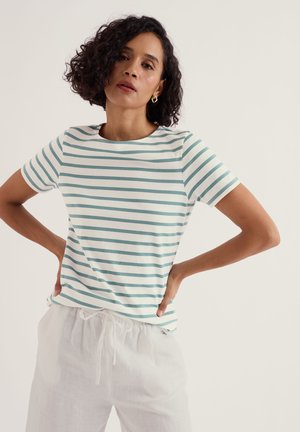 Femeie cu păr scurt și creț, purtând cercei rotunzi de aur, o bluză cu mânecă scurtă cu dungi albe și verzi, și pantaloni albi cu șnur, cu mâinile pe șolduri.