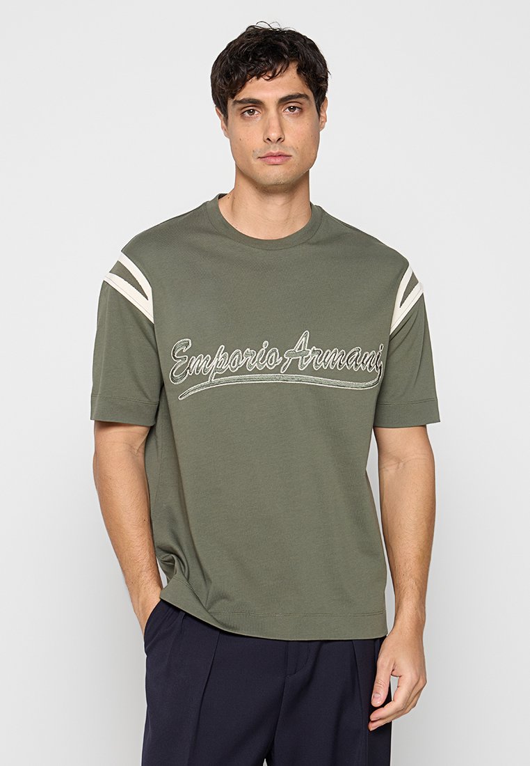 Hombre con camisa verde oliva de manga corta con logo blanco "Emporio Armani" y rayas blancas en los hombros, combinado con pantalones oscuros.
