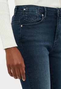 Σκούρο μπλε τζιν παντελόνι με εφαρμοστή γραμμή, ψηλή μέση και μπροστινές τσέπες. Λείος υφασμάτινος σχεδιασμός με ορατές ραφές και ασημένια μεταλλικά στοιχεία.